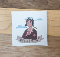 On white sticker paper, close-up image of cartoon illustrated Amelia Earhart wearing her flight goggles on her forehead, rose-coloured scarf and brown leather bomber jacket. Two light blue clouds in the background. Below her image is a light brown ribbon bearing her name. Sticker measures 2 by 2 inches in area.