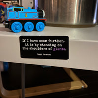 Picture of two by three inch black rectangular fridge magnet with quote by Isaac Newton reading "If I have seen further, it is by standing on the shoulders of giants", on the side of a metal end table with children's toys in the fore and backgrounds.