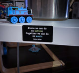 Picture of two by three inch black rectangular fridge magnet with quote by Helen Keller reading "Alone we can do so little; Together we can do so much", on the side of a metal end table with children's toys in the fore and backgrounds.