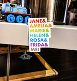 Picture of two by three inch white rectangular fridge magnet with the all caps names of Jane, Amelia, Marie, Helen, Rosa, Frida and Me stacked vertically. Each name is in a different colour of typeface - in descending order these colours are red, orange, green, turquoise, blue, purple and charcoal grey. This magnet sits proudly on the side of a metal end table with children's toys in the fore and backgrounds.