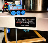 Picture of two by three inch black rectangular fridge magnet with quote by Shirley Chisholm reading "If they don't give you a seat at the table, bring a folding chair", on the side of a metal end table with children's toys in the fore and backgrounds.